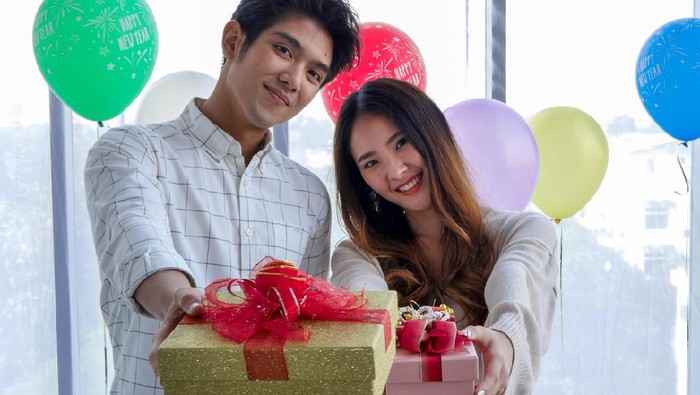 Beautiful Girl Surprise Her Boyfriend With Gift Celebrating Anniversary While He Is Sitting on the Sofa in the Living Room. Romantic Surprise for Husband Boyfriend, Happy Birthday Concept