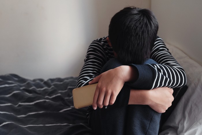 Young Asian preteen teenager boy hugging his knee in his bedroom with smartphone, Cyber bullying in kid, depressed child mental health