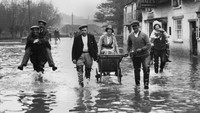 Potret seorang wanita yang menaiki gerobak semen saat tengah dievakuasi dari banjir yang merendam kawasan Bampton pada tahun 1932. Ada pula warga yang digendong agar tak basah terendam banjir. Fox Photos/Getty Images.