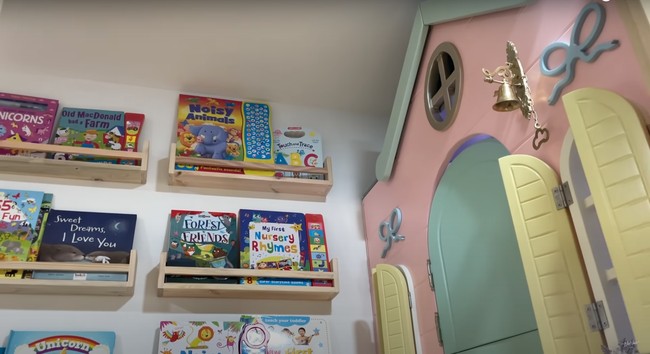 Pada bagian salah satu dindingnya terdapat rak-rak yang digunakan untuk meletakkan buku-buku. Tepat di sampingnya terdapat rumah-rumahan yang dilengkapi dengan lonceng kecil di bagian depannya. Foto: YouTube/NisNaz Channel