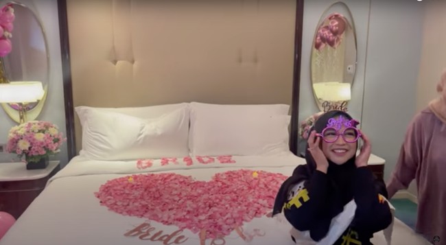 Kamar hotel tersebut tampak sudah didekorasi sedemikian rupa oleh para sahabat Ria Ricis. Kasurnya pun telah dihias dengan taburan bunga yang dibentuk menjadi tulisan bride dan bentuk hati. Foto: YouTube/Tri Atika