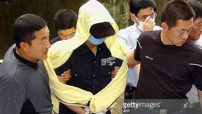 SEOUL, REPUBLIC OF KOREA:  Yoo Young-Chul (C), a 33-year-old former convict wearing a yellow raincoat and a face mask, is handcuffed as he is escorted by police in Seoul, 18 July 2004, to a mountain area where digging is underway for some of the victims he has confessed to killing.  Investigators said Yoo had used an electric saw, knives, hammers, axes and scissors to kill or chop up some 19 South Korea victims -- mostly female who worked as masseuse and hostesses at karaoke bars.  REPUBLIC OF KOREA OUT   AFP PHOTO/DONG-A ILBO  (Photo credit should read AFP/AFP via Getty Images)