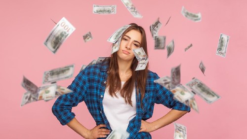 Unhappy girl in checkered shirt holding hands on hips and looking at camera with upset tired expression while money falling around, worried about financial crisis, debts. indoor studio shot isolated