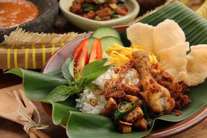 Nasi Uduk, a traditional dish from Betawi, Jakarta. A dish of steamed rice infused with fragrant coconut milk and served with variety of side dishes such as fried chicken, sauteed tempeh, omelet, fresh vegetables, cracker crisp, fried shallots and spicy peanut sauce. All the food items have been arranged on a banana-leaf-lined earthenware plate, ready to be served as a one-dish-meal. A pair of wooden cutleries is placed next to the plate. Further back in blur are the sautéed tempeh and spicy peanut sauce.