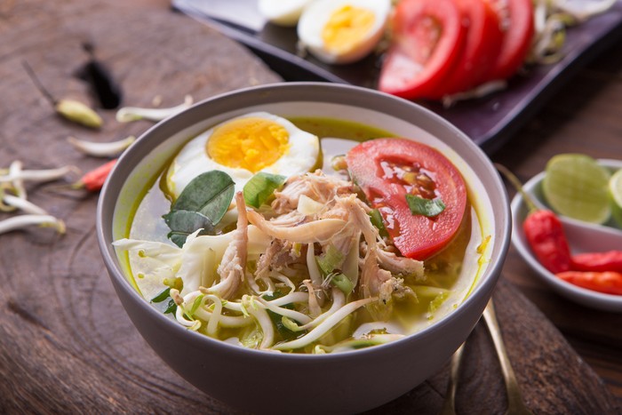 Soto: Indonesian chicken soup with hard boiled eggs, tomato, bean sprout, lemon, cayenne pepper on rustic brown wooden background