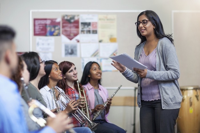 Smiling Indian female tutor holding sheet music while teaching students. Teenage boys and girls are learning from teacher. They are in classroom at high school.