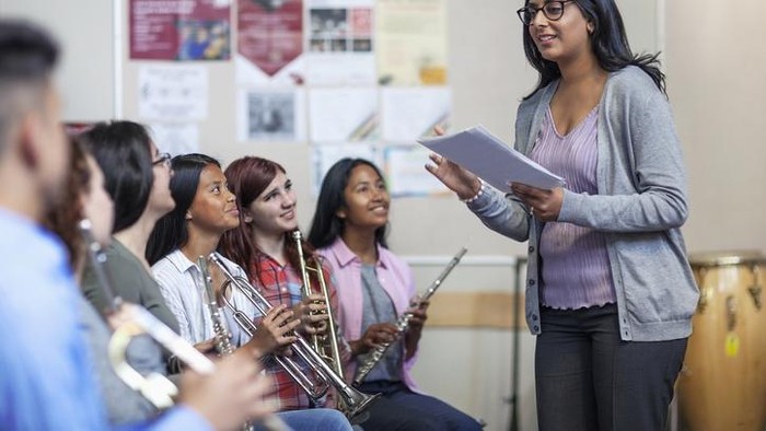 Smiling Indian female tutor holding sheet music while teaching students. Teenage boys and girls are learning from teacher. They are in classroom at high school.