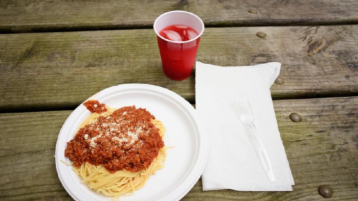 Photograph of a paper plate full of spaghetti with meat sauce and parmeson cheese meal with plastic cup on picnic table.  Cup full of red drink and paper towel with plastic fork on table.