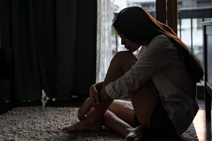 1173052440 Selective focus loneliness young asian woman sitting on bedroom floor near the balcony. Depression sadness breaking up asian teenage girl sitting alone hugging knees closing eyes and thinking.