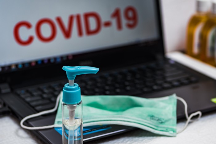 Hand sanitizer bottle and surgical mask on a laptop keyboard that reads COVID-19 on the screen. The concept of preventing office employees from coronavirus disease pandemic.