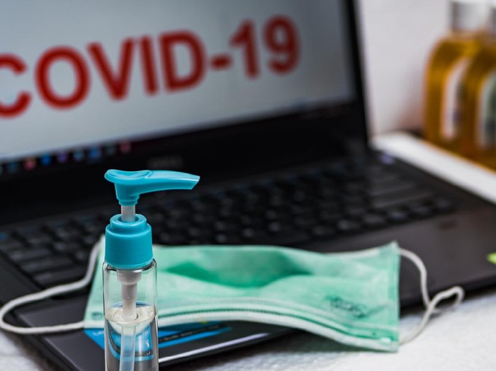 Hand sanitizer bottle and surgical mask on a laptop keyboard that reads COVID-19 on the screen. The concept of preventing office employees from coronavirus disease pandemic.