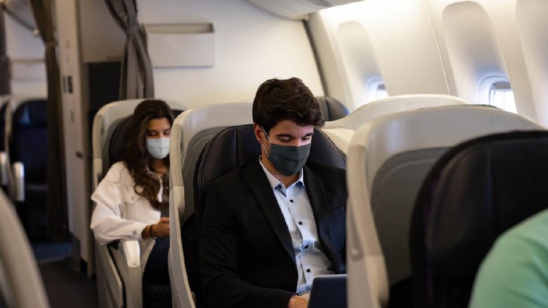 Ilustrasi tempat duduk kelas bisnis di pesawat Business man traveling and wearing a facemask on the plane while using his laptop– COVID-19 pandemic lifestyle concepts