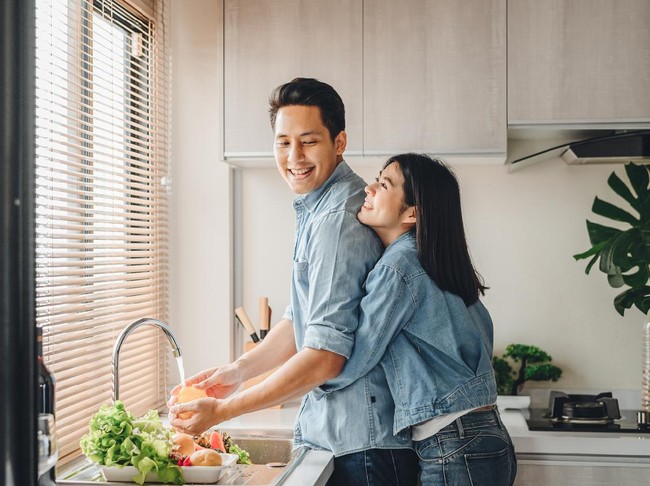 Asian couple lovers hug in the kitchen while cooking at home. A woman hugs a man who is washing vegetables from the back.