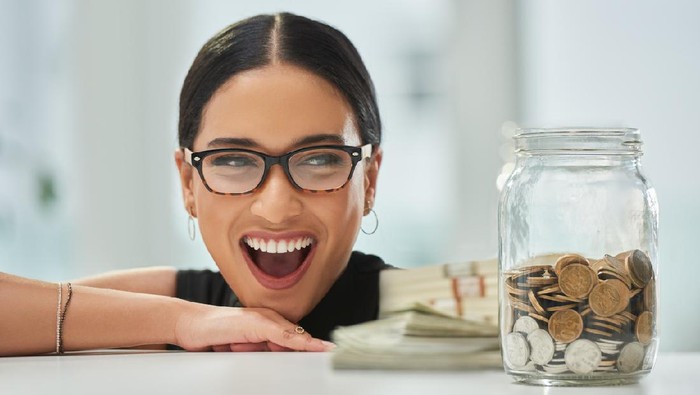 Cropped shot of an attractive young businesswoman looking excitedly over at a jar of coins and wads of cash in her office