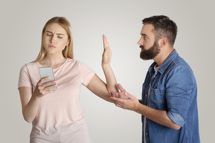 Family problems, indifference and quarrel. Busy young wife looking at phone, typing message and making stop hand gesture for husband, want to talk isolated on gray background, free space, studio shot