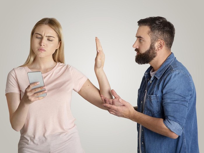 Family problems, indifference and quarrel. Busy young wife looking at phone, typing message and making stop hand gesture for husband, want to talk isolated on gray background, free space, studio shot