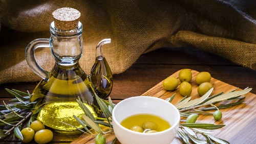 Front view of an olive oil bottle surrounded by green olives and a olive wooden cutting board with more green olives, olive branches and a withe bowl filled with three green olives and olive oil on top. Low key DSLR photo taken with Canon EOS 6D Mark II and Canon EF 24-105 mm f/4L