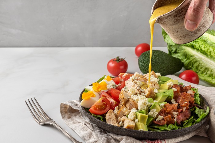 A hand pouring salad dressing into cobb salad with chicken, avocado, bacon, blue cheese, tomato and eggs. Keto diet food. Classic American dish