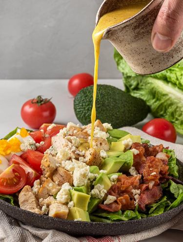A hand pouring salad dressing into cobb salad with chicken, avocado, bacon, blue cheese, tomato and eggs. Keto diet food. Classic American dish