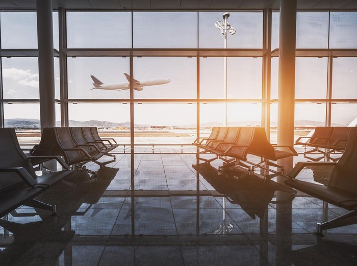Wide-angle view of a modern aircraft gaining the altitude outside the glass window facade of a contemporary waiting hall with multiple rows of seats and reflections indoors of an airport terminal El Prat in Barcelona