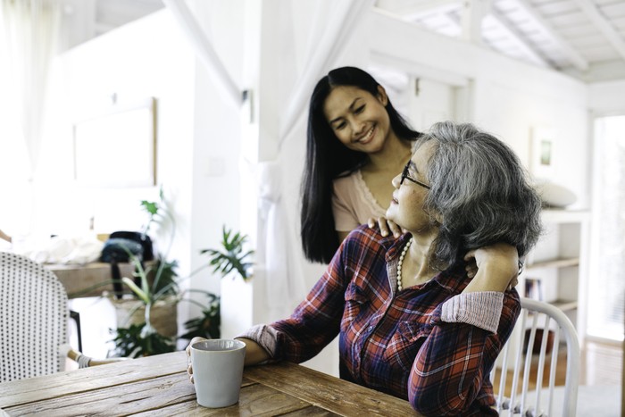 Beautiful adult granddaughter hugging beautiful senior grandmother while sitting on the chair drinking coffee