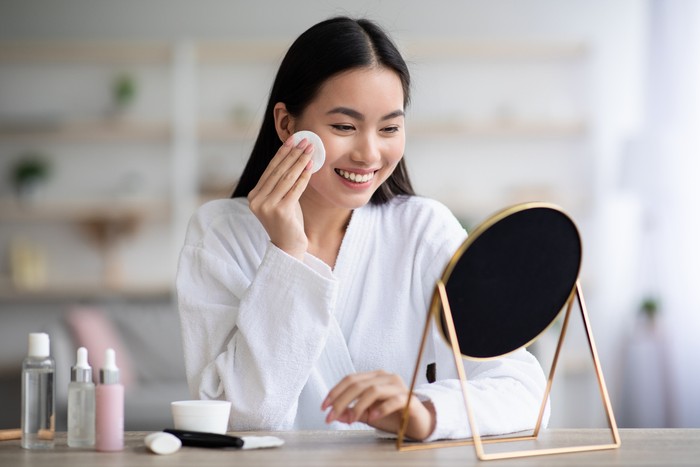 Beautiful asian woman cleaning her face, using cotton pads and cleansing product, looking at mirror in bedroom. Young attractive korean lady using face toner and cotton pad, home interior, empty space