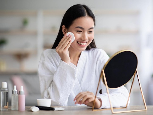 Beautiful asian woman cleaning her face, using cotton pads and cleansing product, looking at mirror in bedroom. Young attractive korean lady using face toner and cotton pad, home interior, empty space