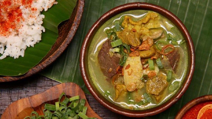Empal Gentong, the signature, traditional beef soup from Cirebon in West Java. Savory coconut milk soup cooked in earthenware pot served with cubes of beef and offal. Topped with condiments of fried shallots and scallions with the red chili powder as extra option. A plate of steamed rice topped with chili powder is served with the soup. The soup and the rice are plated on earthenware crockery; then arranged on a table lined with banana leaf.