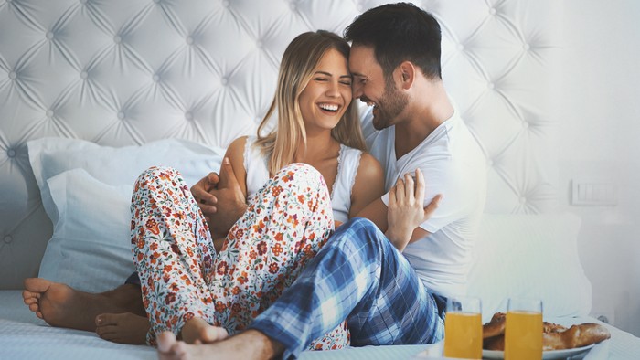 Closeup front view of late 20s couple relaxing in bed and having some juice and bagels. They are laughing and having some light conversation, its weekend morning and they have all the day for themselves.