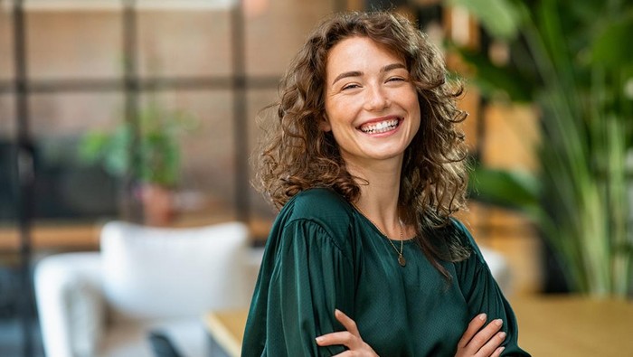 Portrait of young smiling woman looking at camera with crossed arms. Happy girl standing in creative office. Successful businesswoman standing in office with copy space.