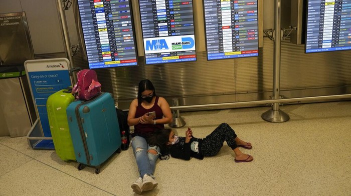 Seorang wanita dan anak-anak dari Brasil menunggu penerbangan mereka di bawah papan yang menunjukkan sekitar selusin penerbangan yang dibatalkan di antara kedatangan yang dijadwalkan, di Bandara Internasional Miami, Senin, 27 Desember 2021, di Miami. Ribuan penerbangan di seluruh dunia dibatalkan atau ditunda pada hari Senin, karena kekurangan staf maskapai karena penyebaran cepat varian omicron dari COVID-19 terus mengganggu musim perjalanan liburan yang sibuk. (Foto AP/Rebecca Blackwell)