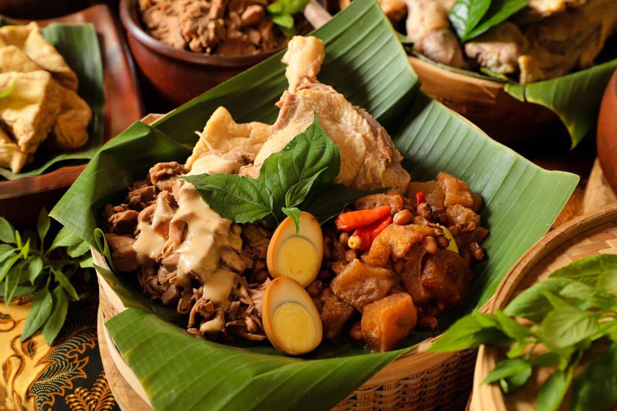 Gudeg Jogja, a Javanesse signature dish of young jackfruit stew; accompanied with white chicken curry and spicy stew of cattle skin cracker. All the dishes are arranged in a bamboo basket as a take-out container. In the background is each of the individual dish that makes up Gudeg Jogja.