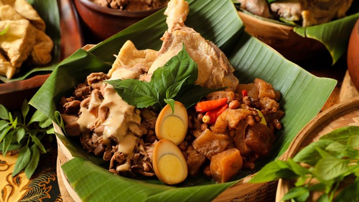 Gudeg Jogja, a Javanesse signature dish of young jackfruit stew; accompanied with white chicken curry and spicy stew of cattle skin cracker. All the dishes are arranged in a bamboo basket as a take-out container. In the background is each of the individual dish that makes up Gudeg Jogja.