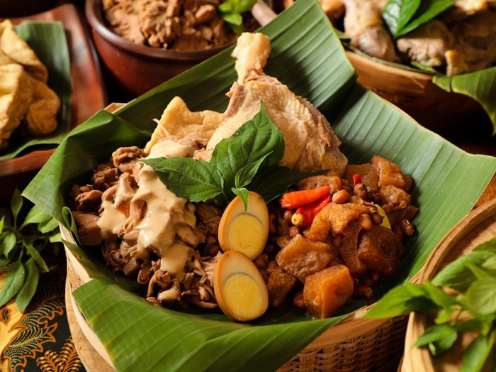 Gudeg Jogja, a Javanesse signature dish of young jackfruit stew; accompanied with white chicken curry and spicy stew of cattle skin cracker. All the dishes are arranged in a bamboo basket as a take-out container. In the background is each of the individual dish that makes up Gudeg Jogja.