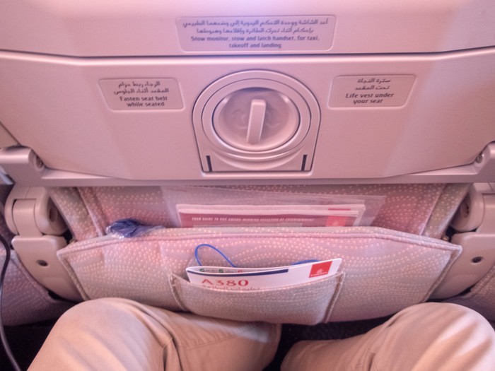 Back of the seat on a commercial long haul flight. This is on an Emirates Airbus A380-800 double decker aircraft and shows the safety sheet in the document pocket in the back of the seat with the passengers legs in the shot.