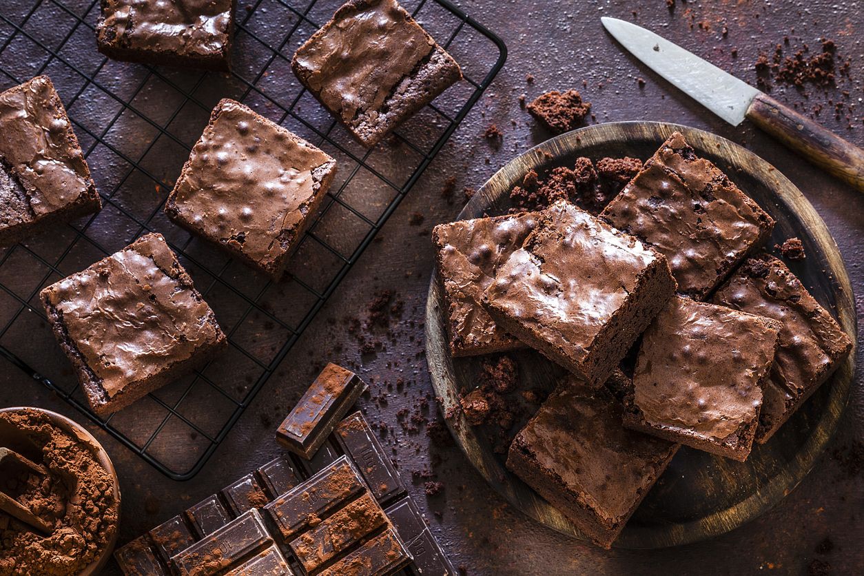 Top view of homemade chocolate brownies shot on dark table. A wooden plate filled with chocolate brownies is at the right and a cooling rack with brownies is at the left. Some chocolate bars are at the bottom. A brown bowl with cocoa powder is at the bottom left corner of an horizontal frame. Predominant color is brown. Low key DSRL studio photo taken with Canon EOS 5D Mk II and Canon EF 100mm f/2.8L Macro IS USM.
