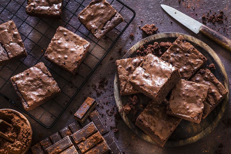 Pemandangan atas brownies coklat buatan sendiri yang diambil di atas meja gelap.  Piring kayu berisi brownies coklat di sebelah kanan dan rak pendingin berisi brownies di sebelah kiri.  Beberapa batang coklat ada di bagian bawah.  Semangkuk coklat dengan bubuk coklat terletak di pojok kiri bawah bingkai horizontal.  Warna utamanya coklat.  Foto studio DSRL low-key diambil dengan Canon EOS 5D Mk II dan Canon EF 100mm f/2.8L Macro IS USM.