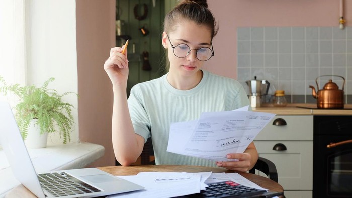 Indoor picture of young European female sitting at home at table reading data in sheets of paper with pencil in order to figure out something important and check it, feeling relaxed and confident