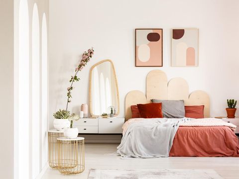 Real photo of a wide bed standing between a shelf with a plant and  a shelf with a mirror in a rusty bedroom interior with two posters on a wall and two gold, small tables with plants