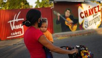 Perempuan menggendong anaknya melintas di depan mural Presiden Hugo Chavez, di kampung halaman Chavez di Sabaneta, Barinas (7/1/2022). Hugo Chavez menjabat sebagai presiden sejak 2002 hingga ia meninggal dunia karena sakit pada 2013. 
