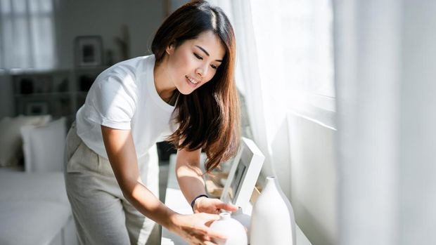 Young Asian woman enjoys her time at home, decorating and organising picture frames and vases on the shelf by the window