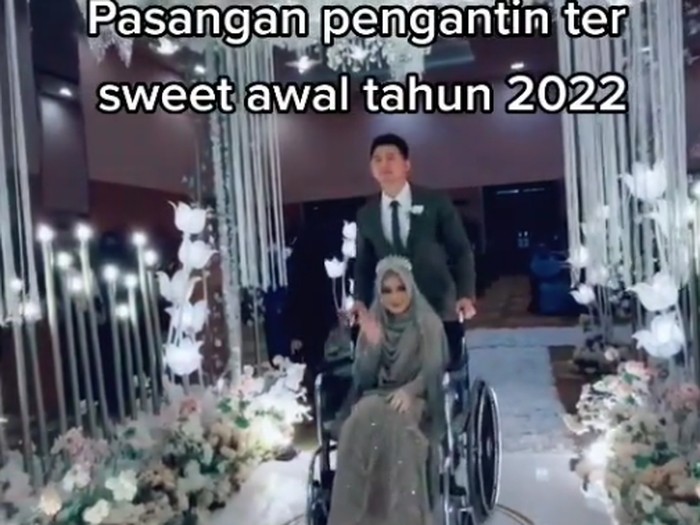 Foto pengantin pria mendampingi pengantin wanita yang memakai kursi roda.