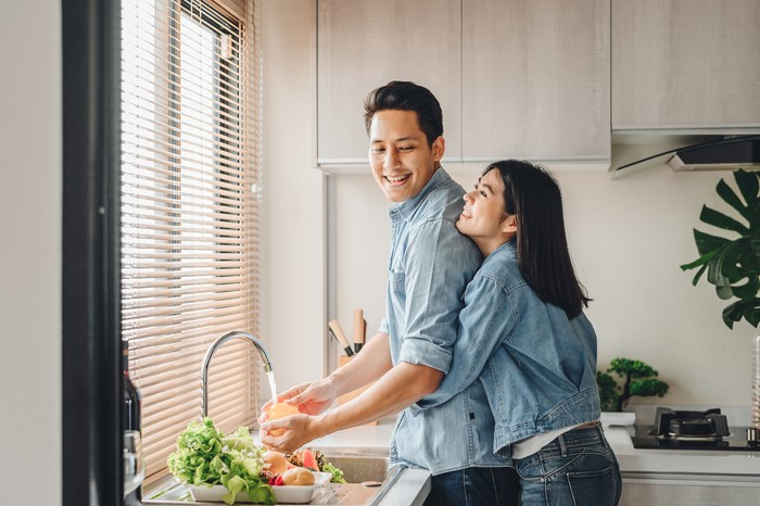 Asian couple lovers hug in the kitchen while cooking at home. A woman hugs a man who is washing vegetables from the back.