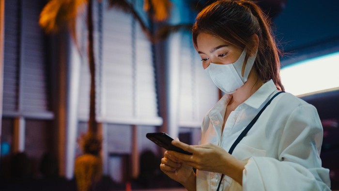 Young Asia businesswoman in fashion office clothes wearing medical face mask using smart phone typing text message while stand outdoors in urban modern city at night. Business on the go concept.