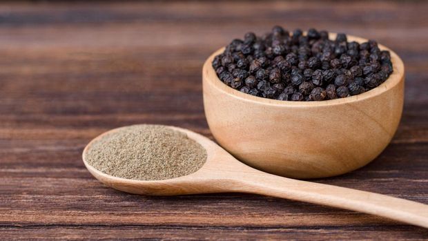 Black pepper seeds or peppercorns ( dried seeds of piper nigrum ) in wooden bowl and spoon isolated on wood table background.