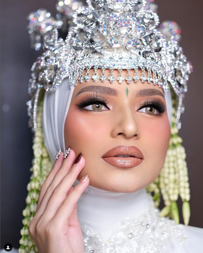 Selain Hollywood Glam, tema Glamour Look juga bisa jadi inspirasi rias pengantin hijab. MUA Nanathnadia, mengaplikasikan tema Glamour Look untuk pengantin wanita berhijab yang mengenakan siger Sunda. Kunci dari tampilan ini adalah gradasi alis yang terlihat natural dan lipstick kombinasi nude gloss. Hijab putih yang dikenakan oleh pengantin wanita semakin menambah kesan flawless dan cantik. Foto: Dok. Instagram @nanathnadia.