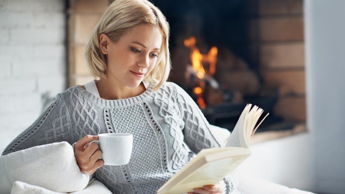 A beautiful young woman reading a book and enjoying a warm beverage near a fireplace