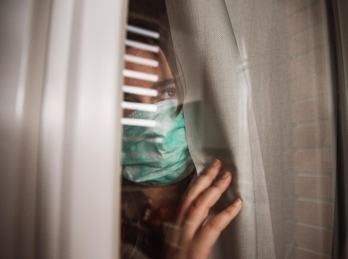 A young woman in quarantine wearing a mask and looking through the window