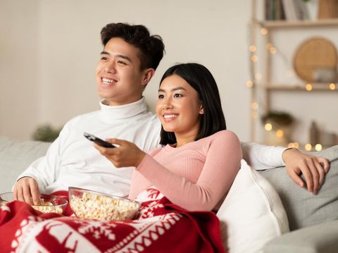 Asian Spouses Enjoying Christmas Eve Watching TV And Xmas Movies Sitting On Couch In Living Room At Home. Asian Family Watches Television Indoors. New Year And X-Mas Celebration Concept