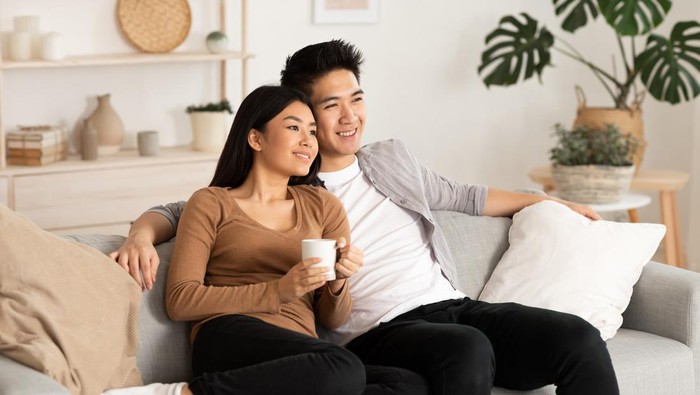 Asian Spouses Enjoying Christmas Eve Watching TV And Xmas Movies Sitting On Couch In Living Room At Home. Asian Family Watches Television Indoors. New Year And X-Mas Celebration Concept